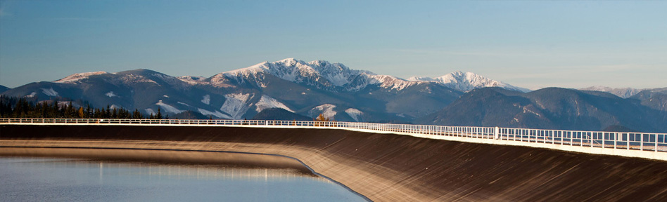 Obrázek zobrazuje přehradu rozprostírající se nad vodní plochou se zasněženými horami v pozadí. Scéna zobrazuje přehradu patřící společnosti Slovenské elektrárne