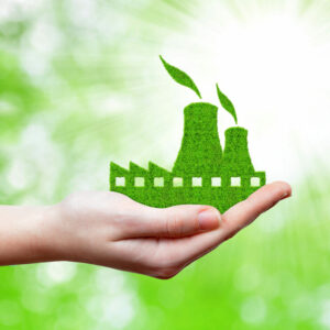 Ruka držící model továrny, která je vyrobena z trávy. Pozadí je zelené s rozmazanými světly, což evokuje ekologii a udržitelnost. Z komínů továrny rostou zelené listy, což symbolizuje šetrnost k životnímu prostředí.